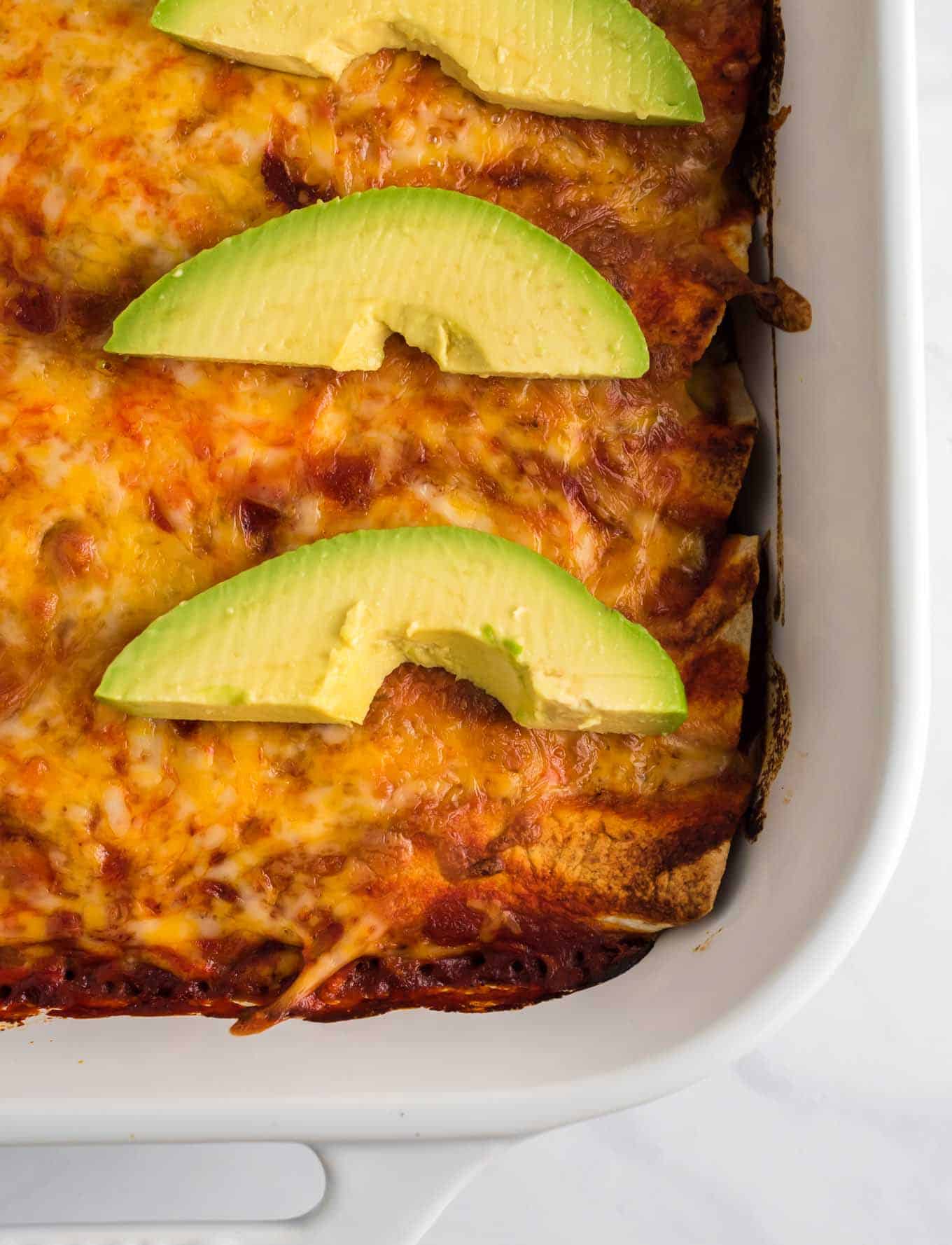 enchiladas in a white pan with sliced avocado on top