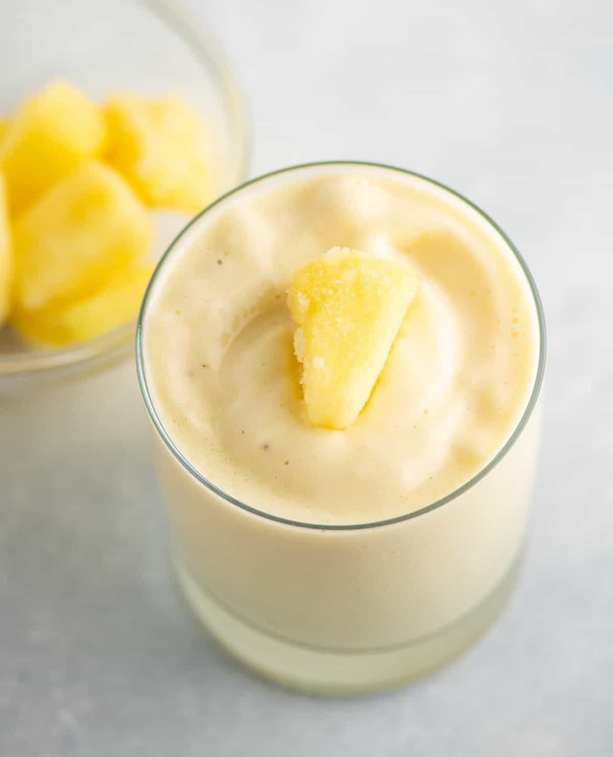 pineapple smoothie in a glass with frozen pineapple on top and frozen pineapple in a bowl in the background