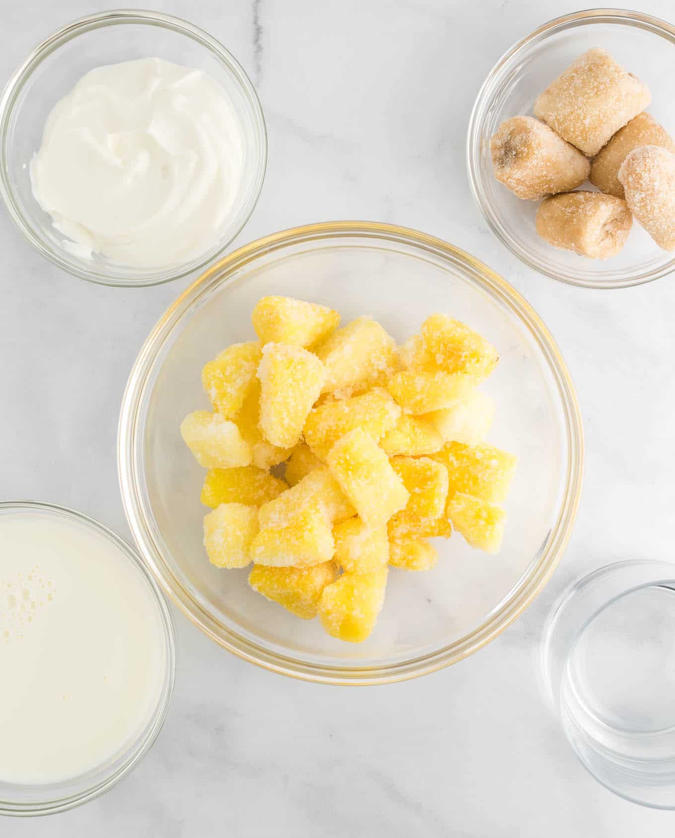 the ingredients in clear bowls - frozen pineapple chunks, greek yogurt, frozen banana chunks, and milk