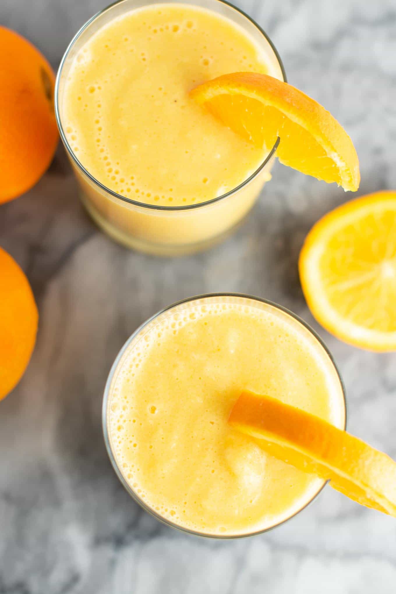 orange smoothie in a clear glass with a slice of orange on the rim from an overhead view with oranges on the side