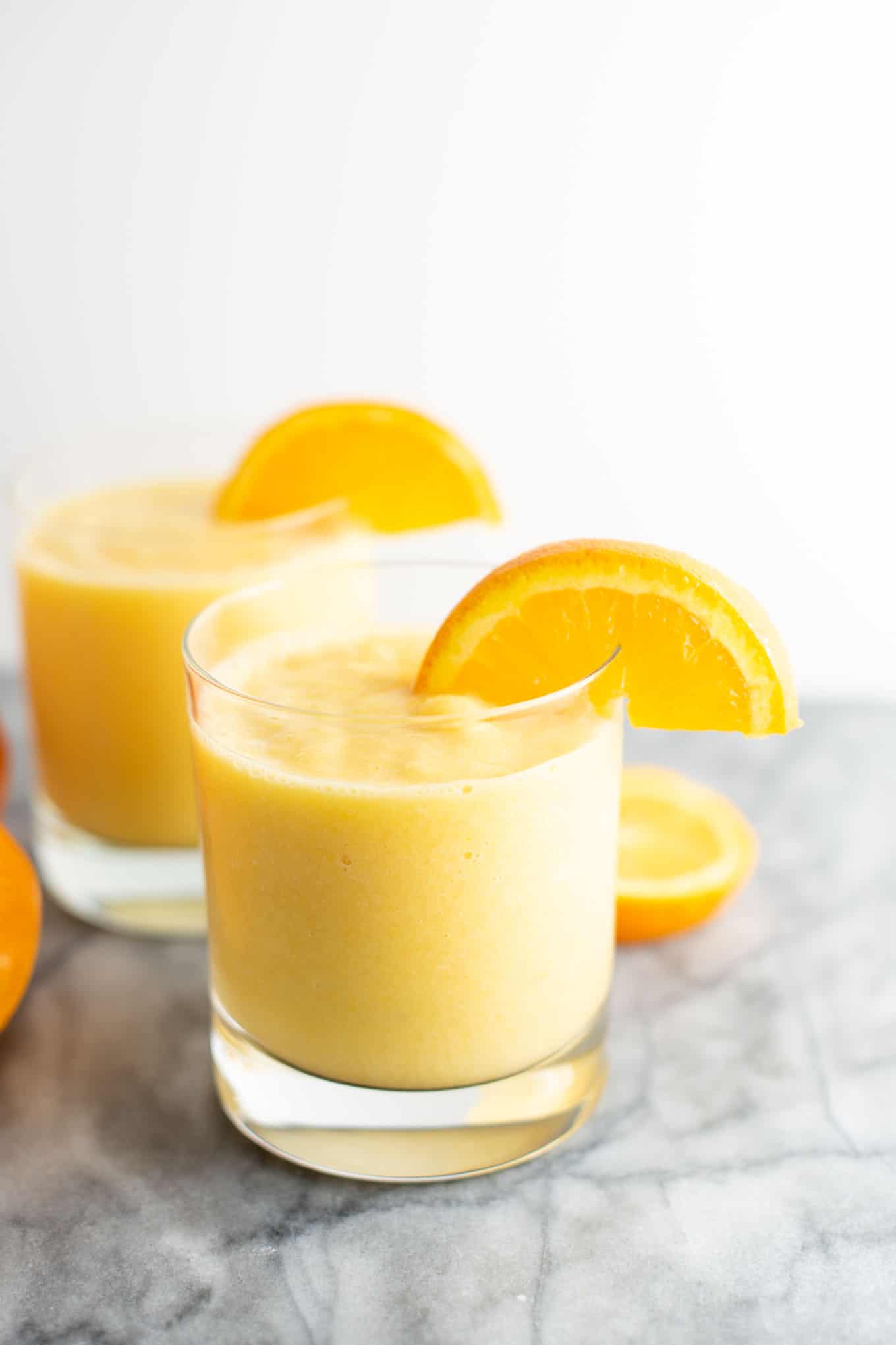 two clear glasses filled with orange smoothie, each with a slice of orange on the rim