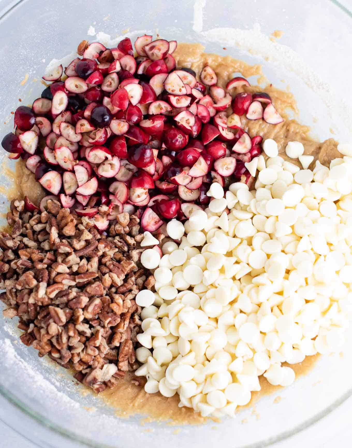 mixing bowl with the batter topped with chopped cranberries, chopped pecans, and white chocolate baking chips