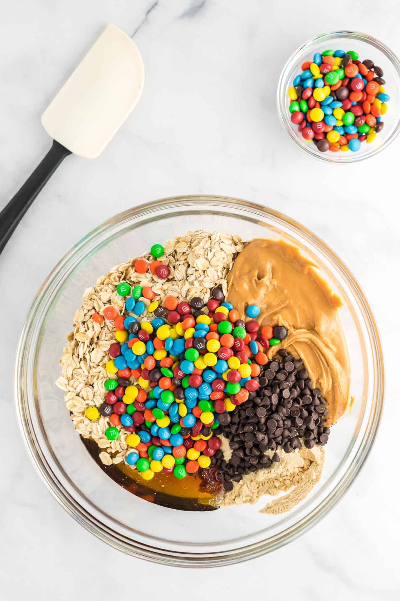 a clear mixing bowl with peanut butter, honey, protein powder, rolled oats, mini chocolate chips, and mini m&m's in it