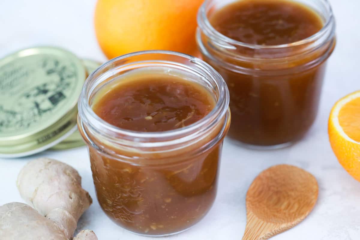 orange sauce in jars with a wooden spoon, ginger, and oranges surrounding them