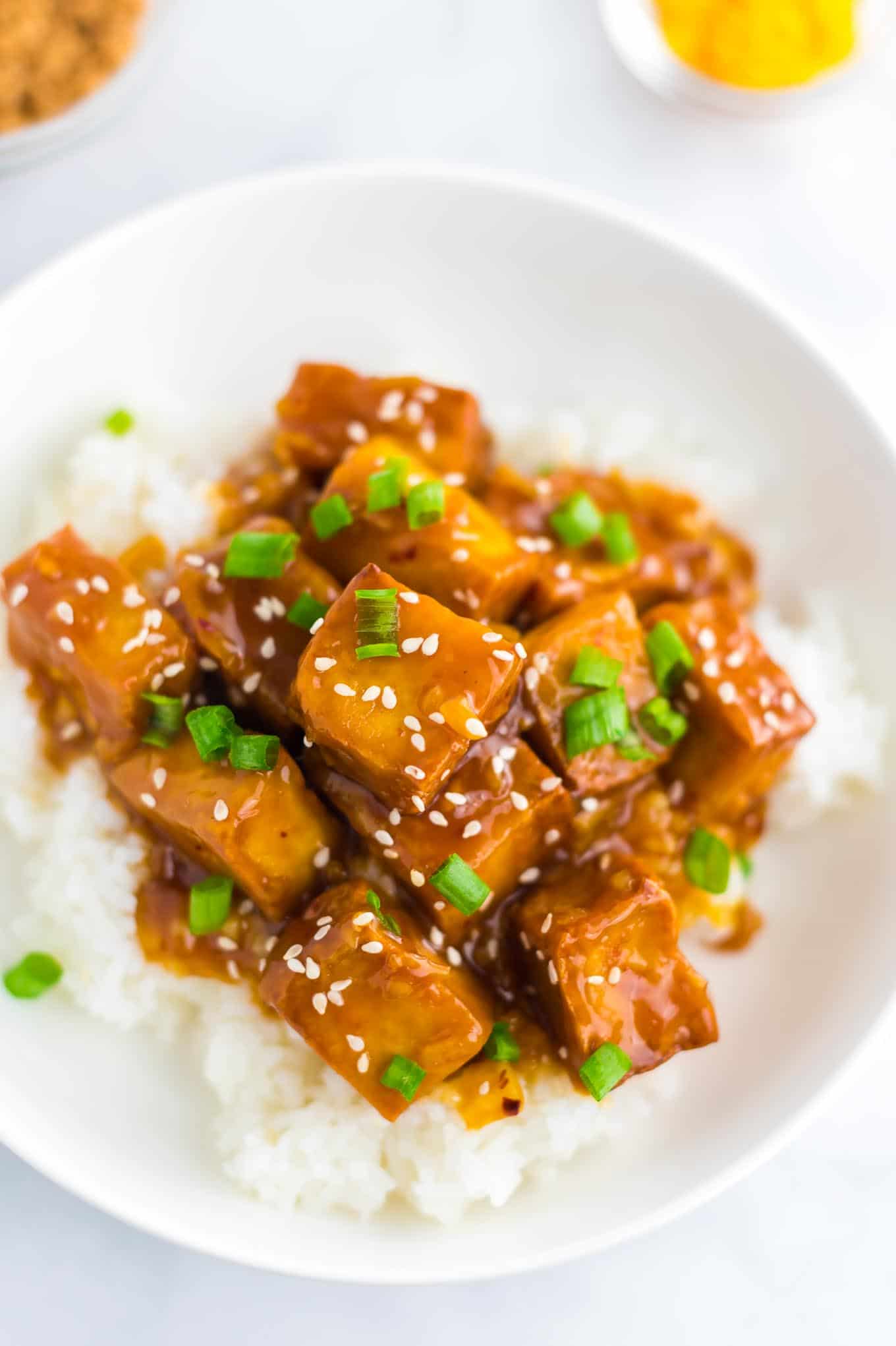 orange tofu over white rice with white sesame seeds and chopped green onions on top