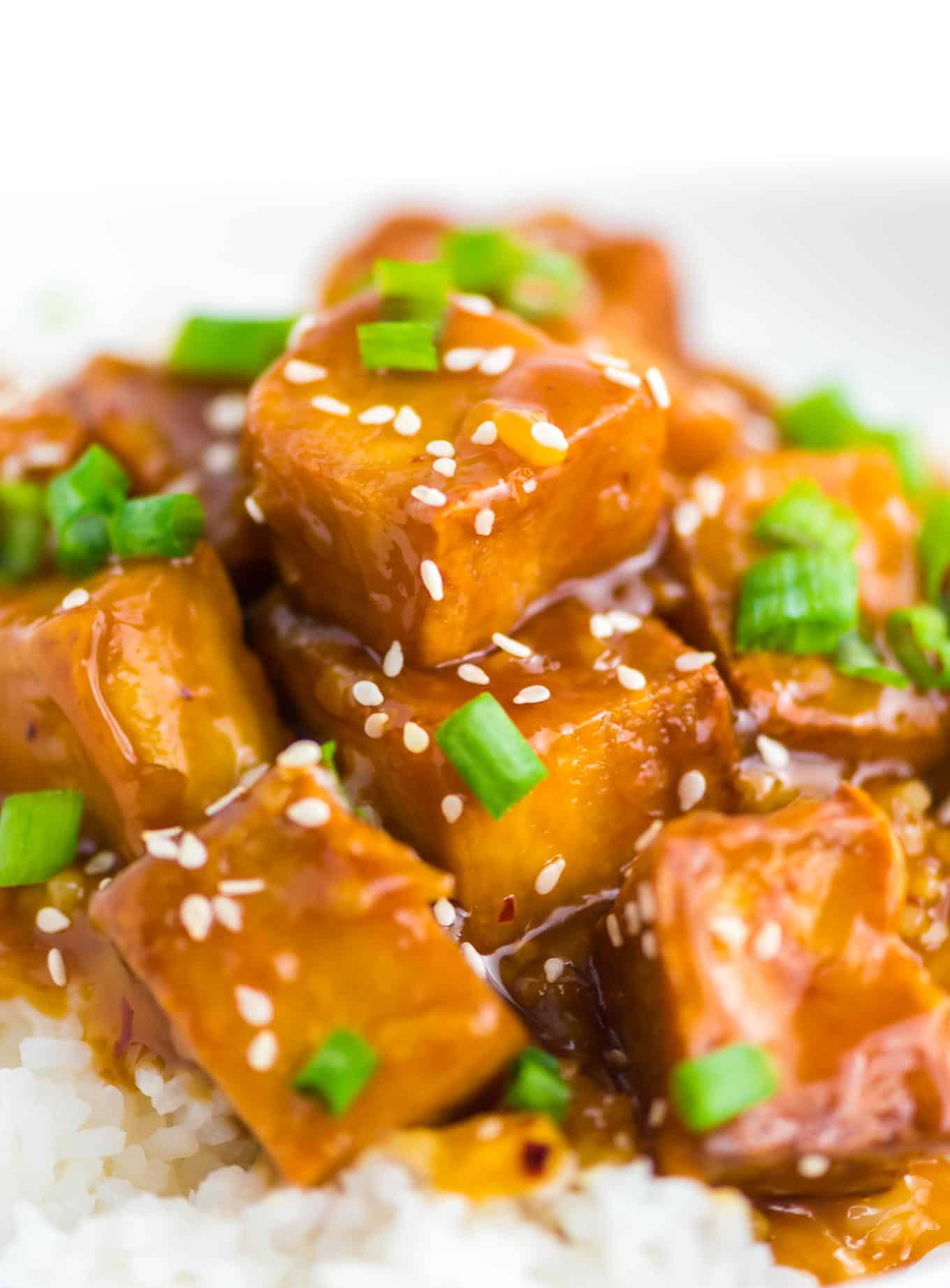 orange tofu over white rice with white sesame seeds and chopped green onions on top