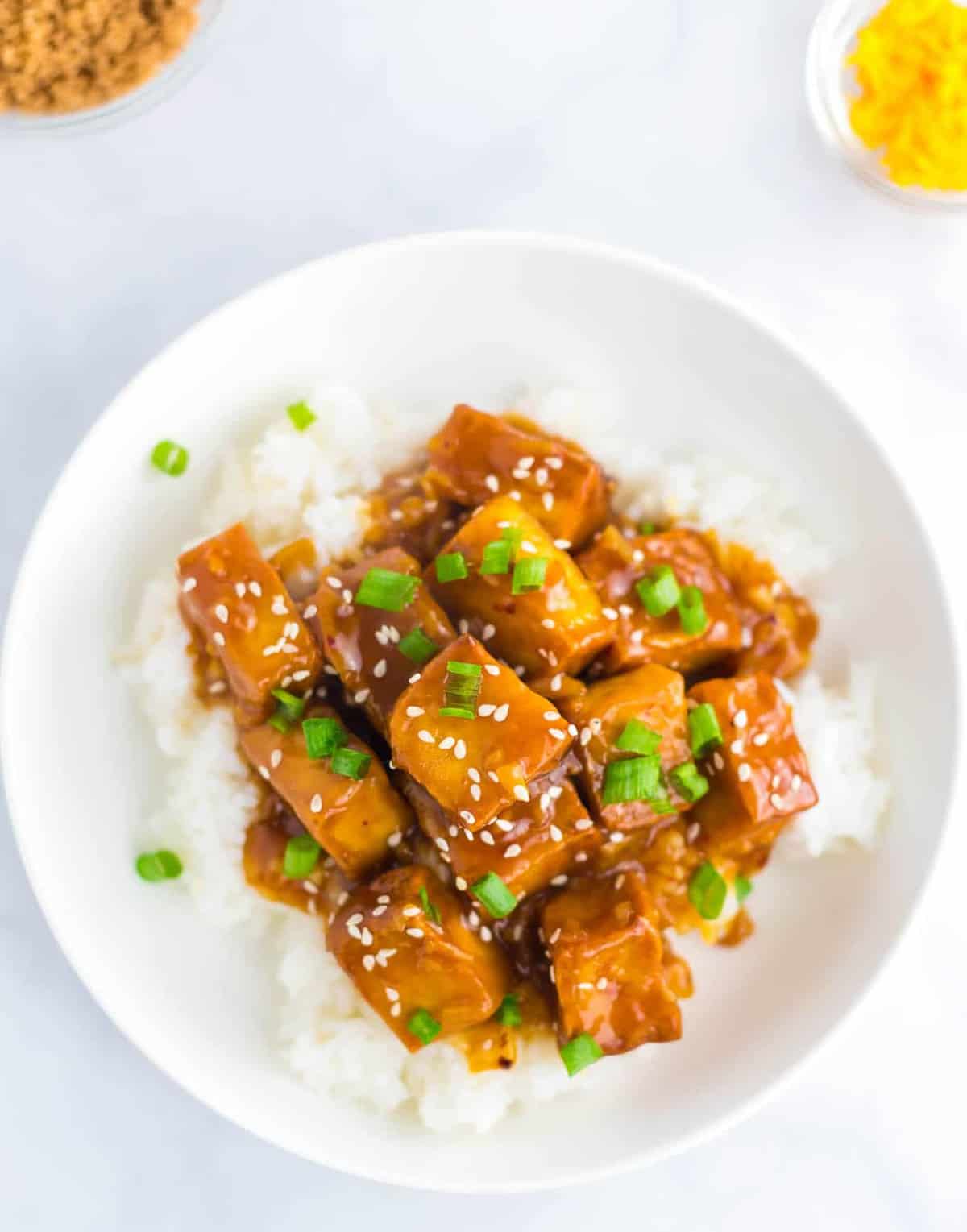 orange tofu in a white bowl with sesame seeds and green onions