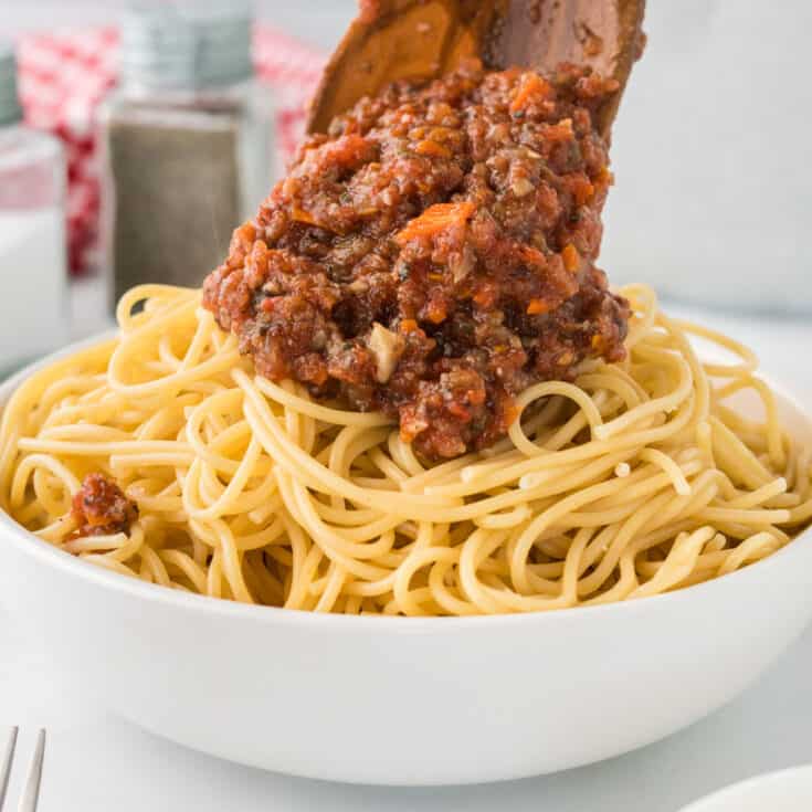 wooden spoon adding bolognese sauce to a bowl of spaghetti noodles
