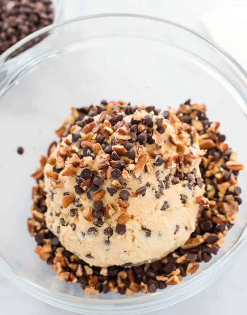 ingredients shaped into a cheese ball being rolled in pecans and mini chocolate chips