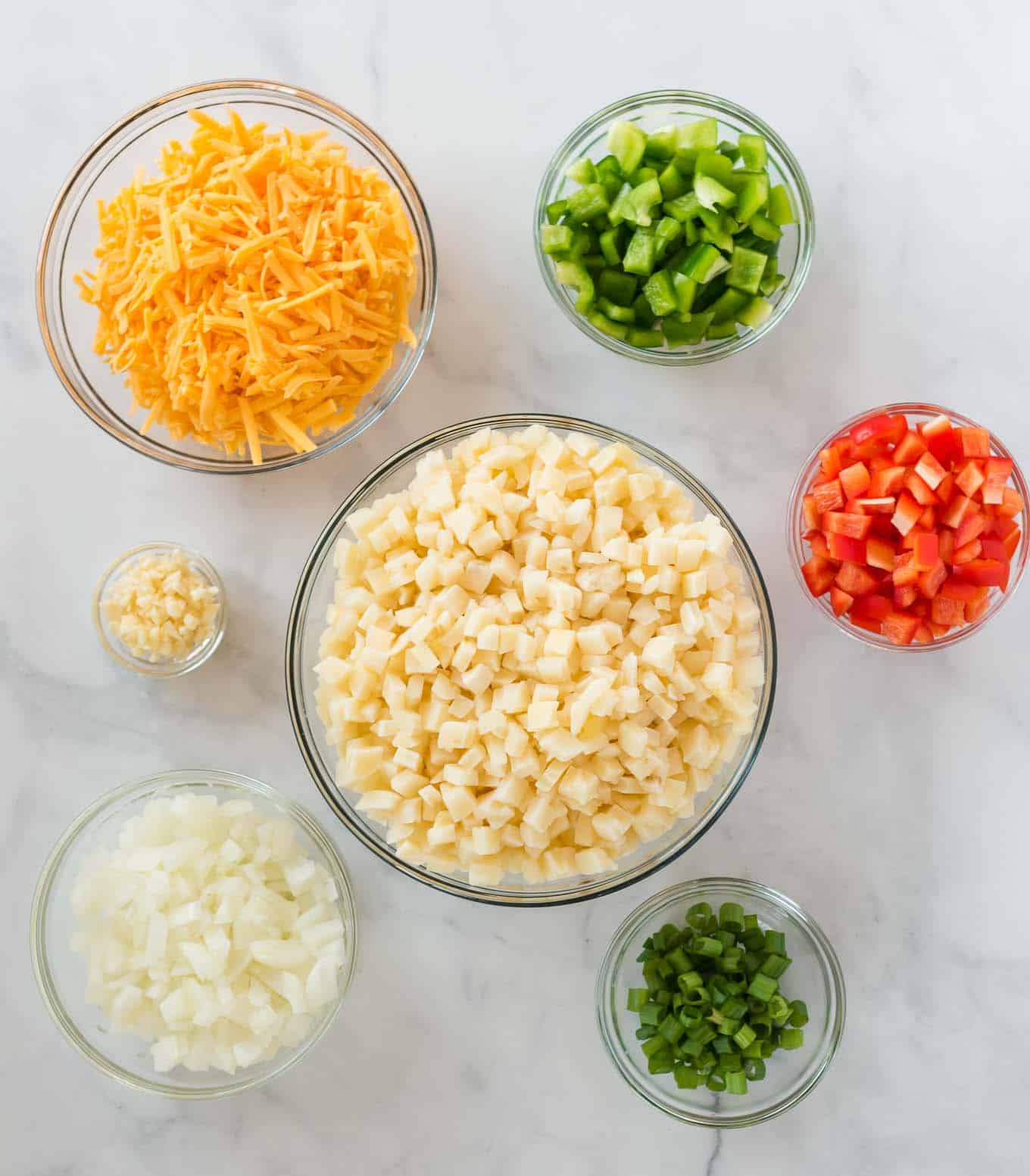 ingredients in separate glass bowls (potatoes, garlic, onion, bell pepper, cheese, green onions)