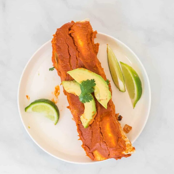 two enchiladas on a white plate topped with avocado and cilantro, with limes on the side