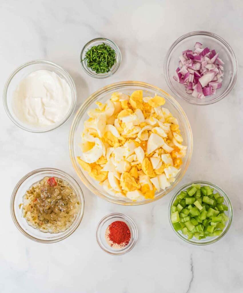 ingredients for egg salad in individual glass bowls - celery, spices, relish, greek yogurt, hard boiled eggs, dill, and red onion