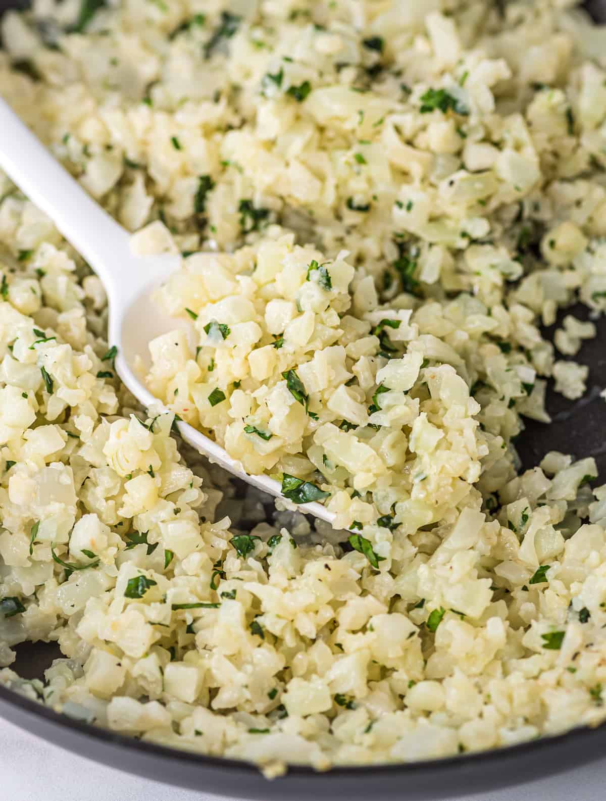 a white spoon taking a scoop of cauliflower rice from the skillet