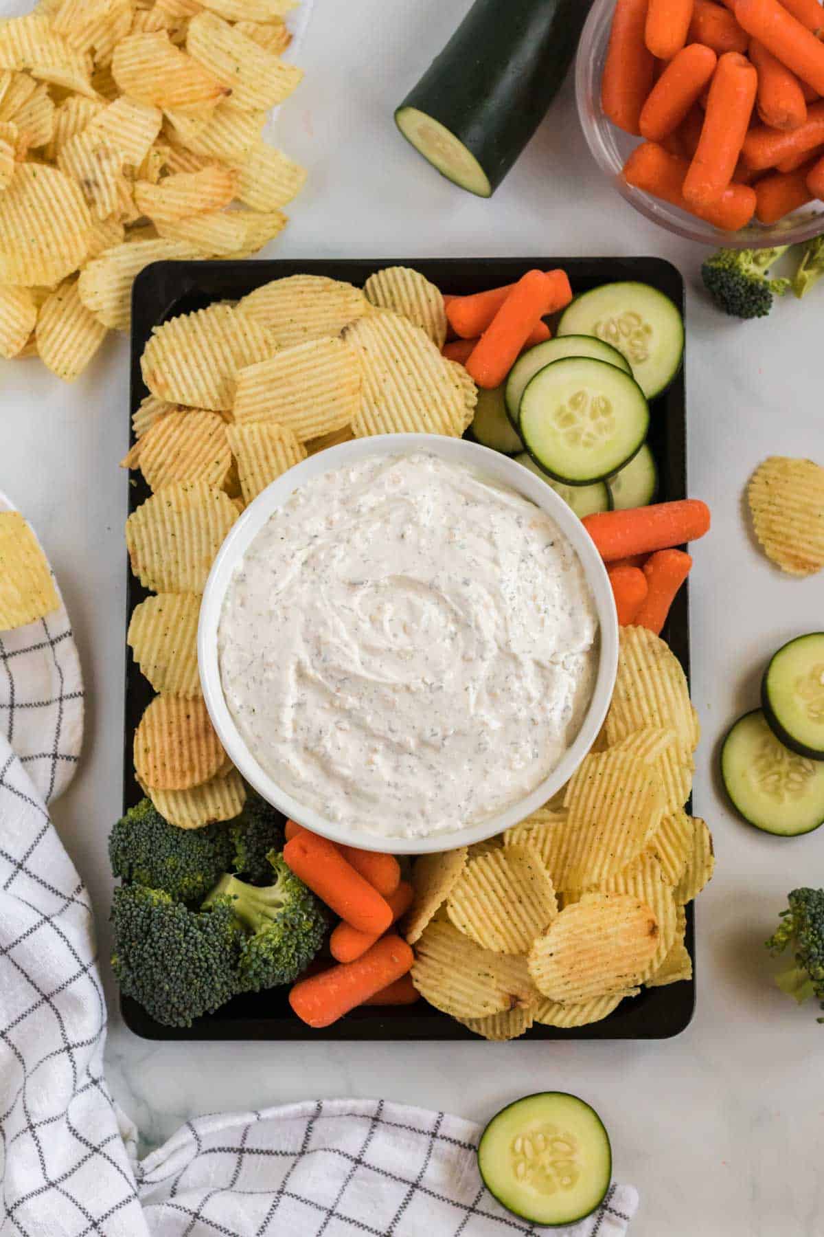 dip in a bowl surrounded by chips, broccoli, baby carrots, and cucumber