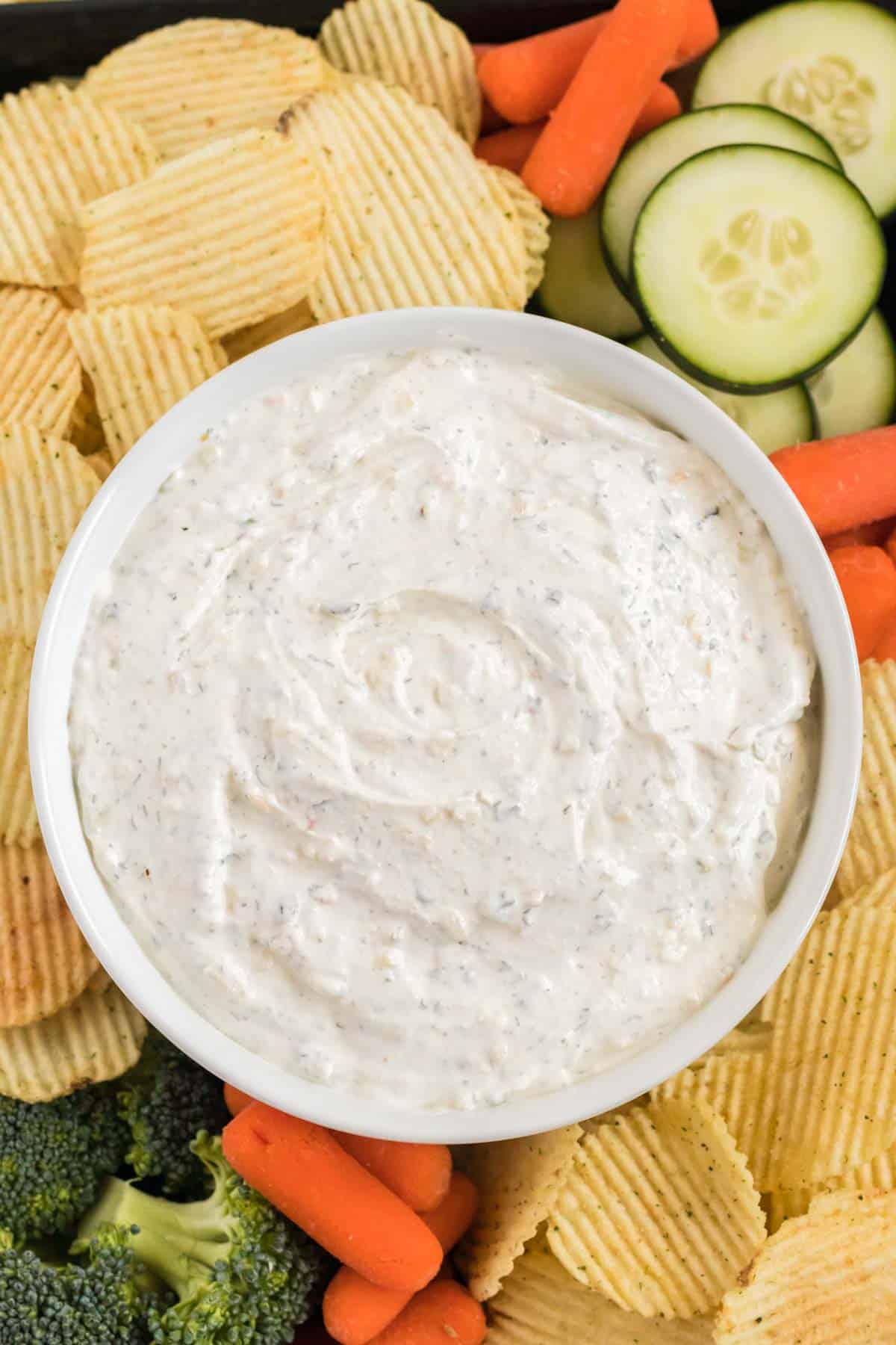 dip in a bowl surrounded by chips, broccoli, baby carrots, and cucumber