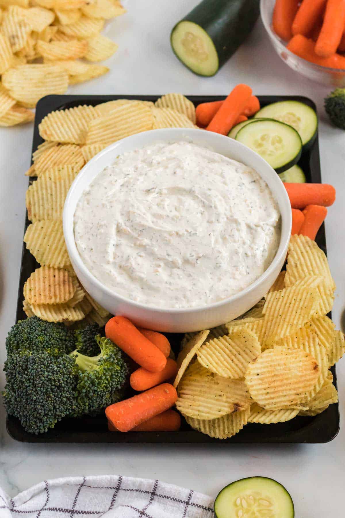 dip in a bowl surrounded by chips, broccoli, baby carrots, and cucumber