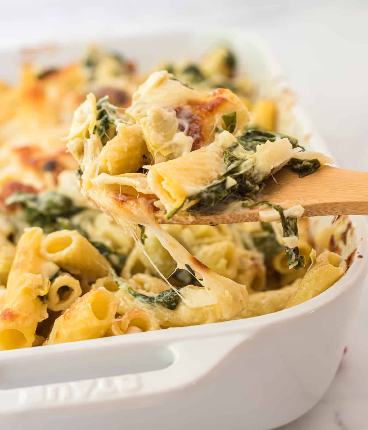 a wooden spoon taking a scoop of spinach artichoke pasta from the baking dish