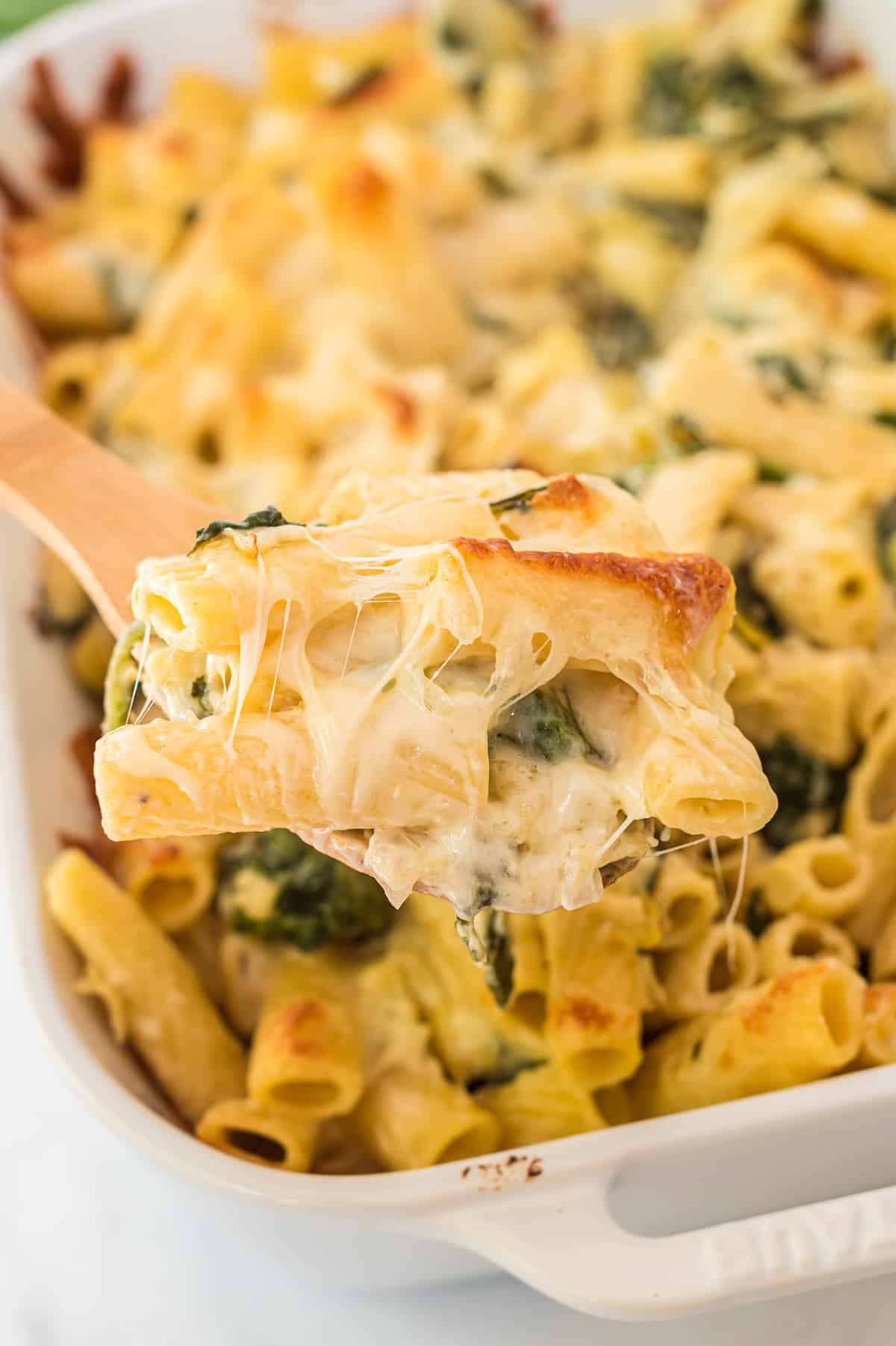 a wooden spoon taking a scoop of spinach artichoke pasta from the baking dish