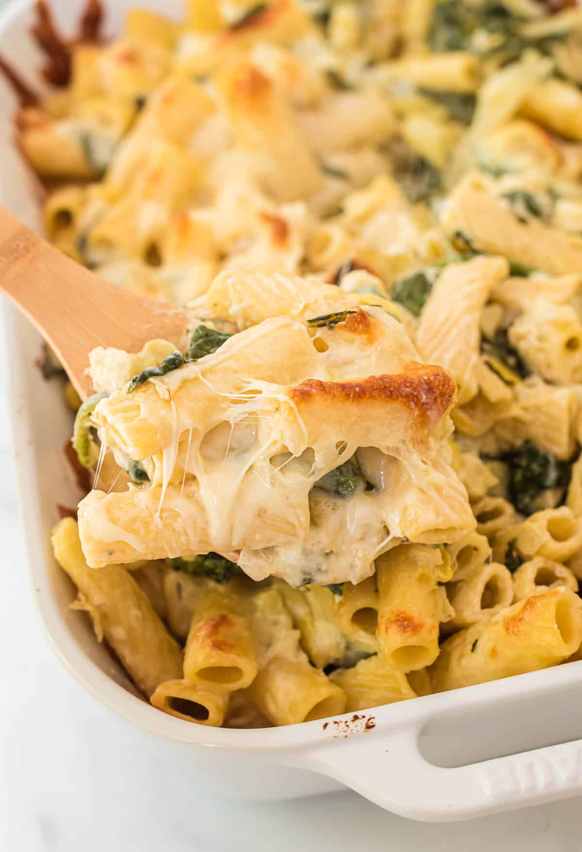 a wooden spoon taking a scoop of spinach artichoke pasta from the baking dish