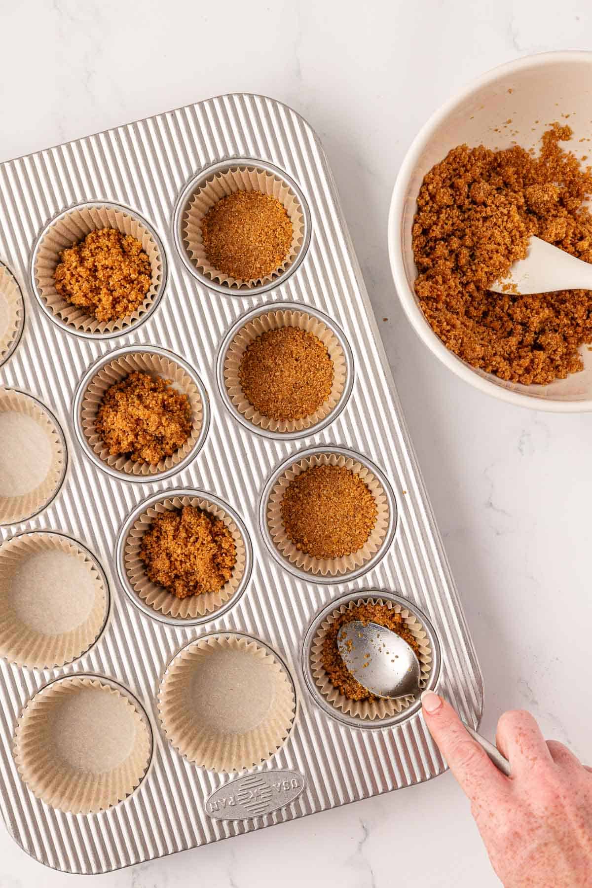 using a spoon to press graham cracker filling into a lined muffin tin